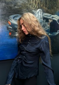 Blusa de seda azul marino con detalle fruncido y acentos de botones, presenta mangas largas y un dobladillo ligeramente ondulado, exhibida sobre un fondo azul.