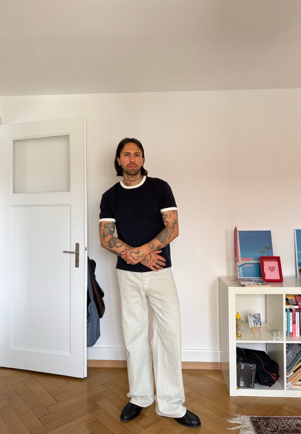 Man met tatoeages draagt een zwart shirt en witte broek en staat in een kamer bij een open deur en een witte plank met foto's en boeken.