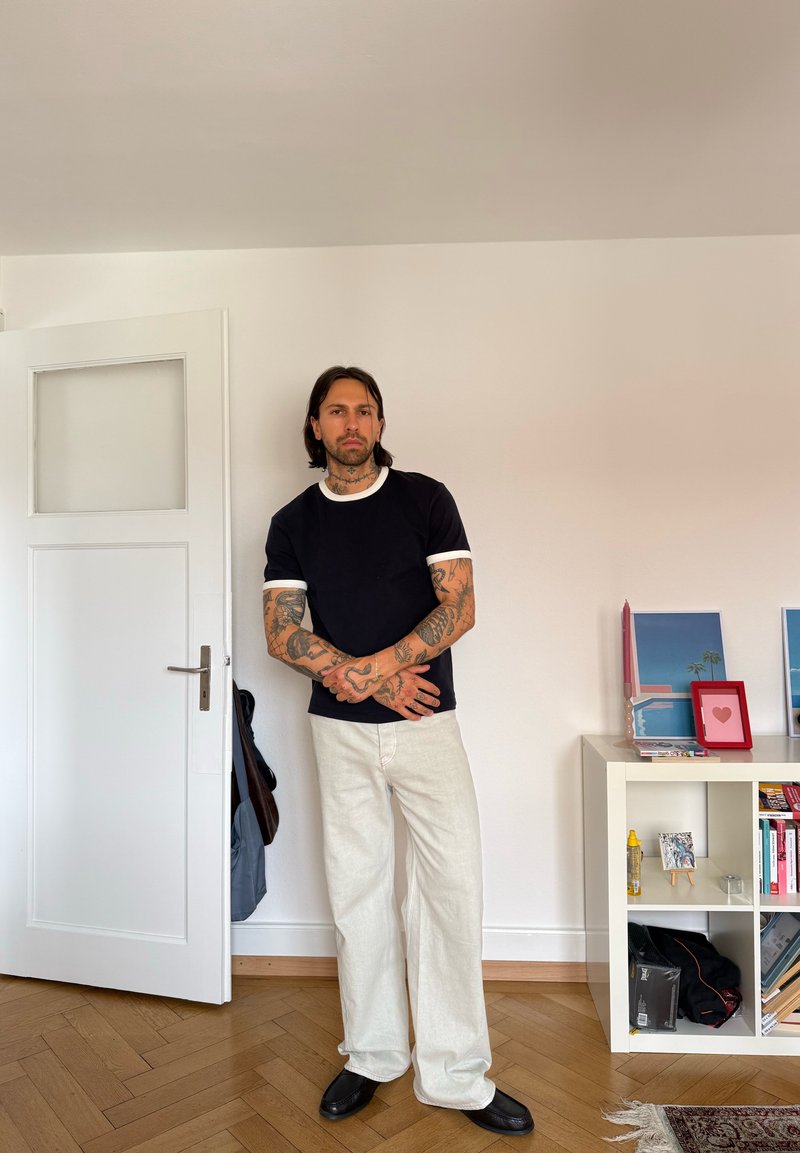 Homme tatoué portant une chemise noire et un pantalon blanc, debout dans une pièce près d'une porte ouverte et d'une étagère blanche avec des photos et des livres.