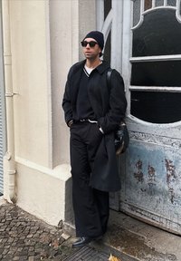 Čierny vlnený kabát cez tmavý sveter, v kombinácii s voľnými čiernymi nohavicami. Doplnky zahŕňajú slnečné okuliare a čiapku. Pozadie z textúrovaného kameňa.