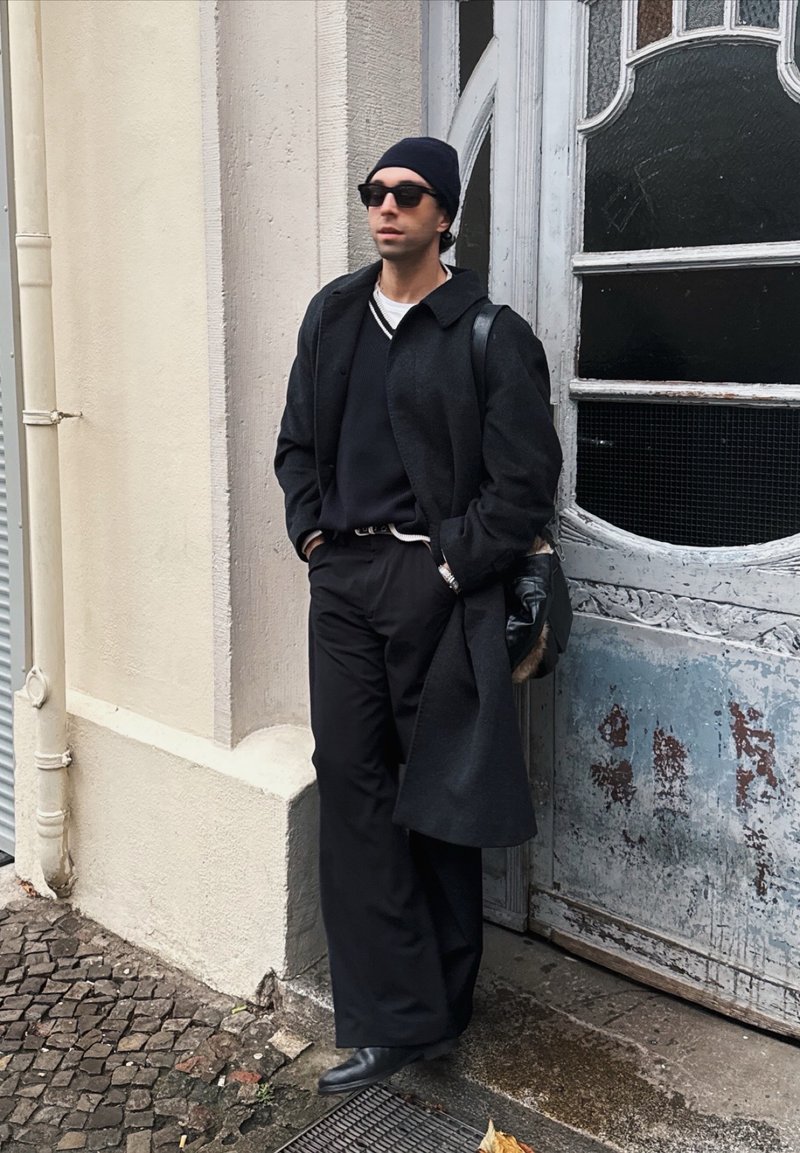 Cappotto di lana nera sopra un maglione scuro, abbinato a pantaloni neri larghi. Accessori includono occhiali da sole e un berretto. Sfondo in pietra lavorata.