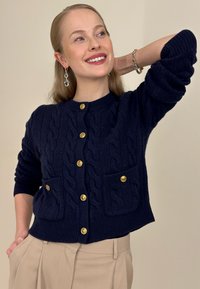 Cardigan en maille bleu marine avec motif torsadé, boutons dorés et deux poches avant. Manches longues et ourlet côtelé.