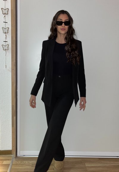 Blazer negro ajustado y pantalones de cintura alta combinados con una blusa negra. Gafas de sol y zapatos de plataforma beige completan el look. Texturas suaves.