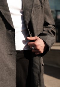 Pelēka, teksturēta mēteļa ar sariem malās nēsāšana virs baltā ribota krekla. Redzamas melnas bikses, uz rokas, kas pieskaras mētelim, ir sudraba gredzens.