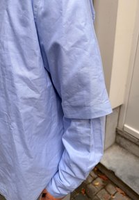 Šviesiai mėlyna ilgomis rankovėmis medvilninė marškinėliai, turintys šiek tiek suraukytą tekstūrą ir atlenktus rankogalius. Matomi siūlių detalės.