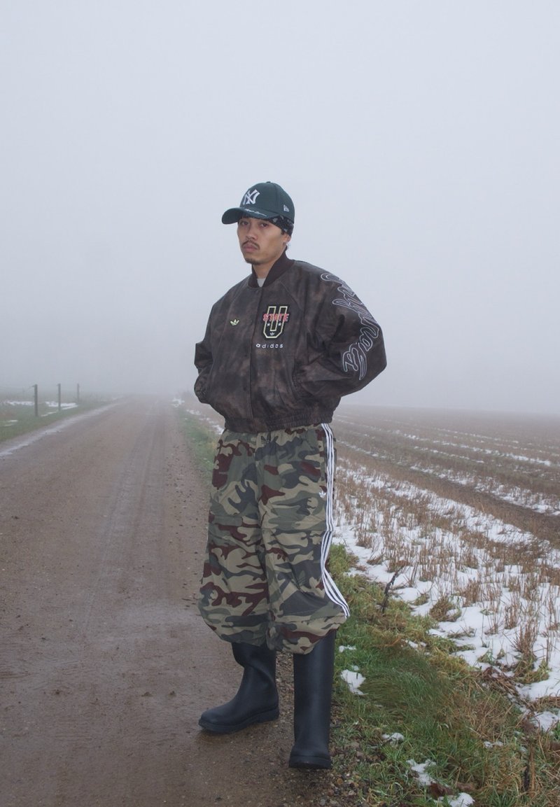 Un homme portant une veste noire, un pantalon camouflage et des bottes en caoutchouc se tient sur une route de terre brumeuse à côté d'un champ enneigé.