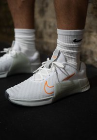 Λευκά αθλητικά παπούτσια με δικτυωτό επάνω μέρος, γκρι λεπτομέρειες, πορτοκαλί λογότυπο Nike και υφασμάτινη σόλα. Φοριούνται με λευκές κάλτσες μέχρι τον αστράγαλο.