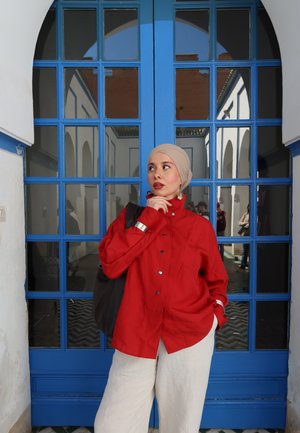 Mujer con pañuelo beige en la cabeza y camisa roja con botones, de pie pensativa con la mano cerca de la barbilla frente a una gran puerta de cristal con cristales azules.
