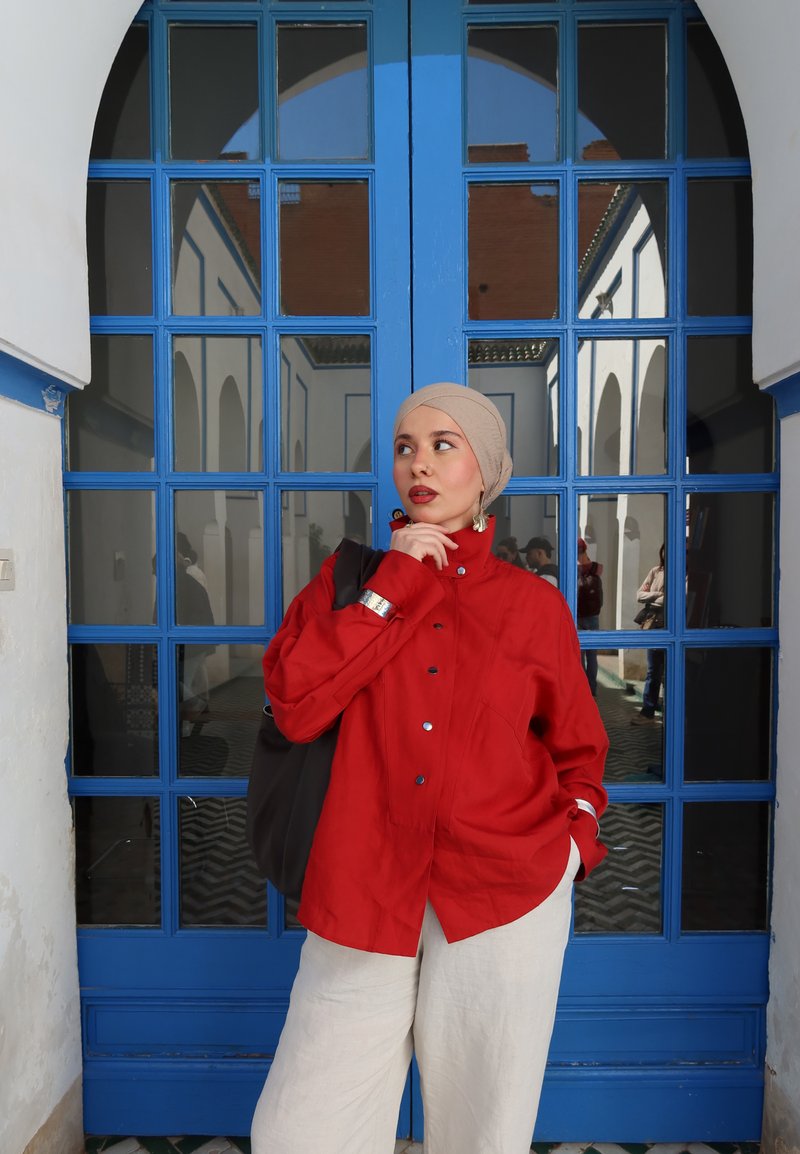 Femme portant un foulard beige et une chemise rouge boutonnée, debout pensivement avec la main près du menton devant une grande porte vitrée aux carreaux bleus.