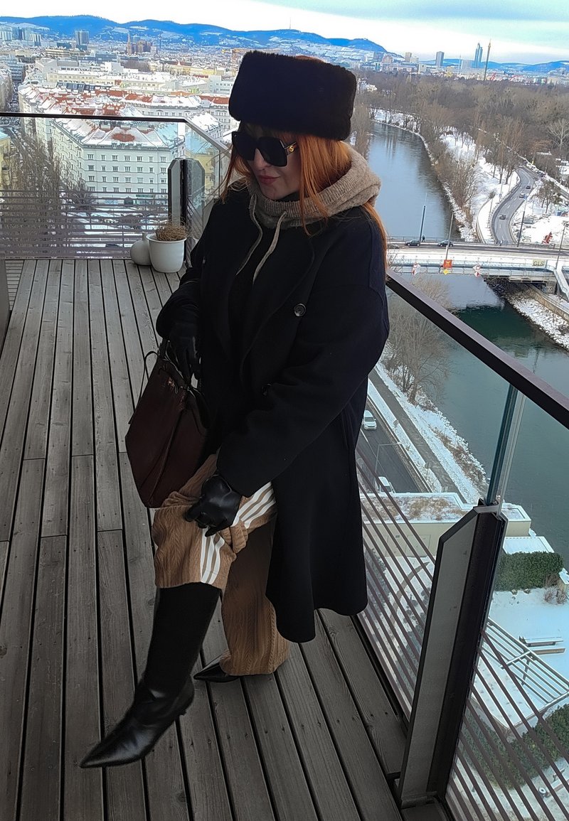 Kobieta w czarnym płaszczu, rękawiczkach i butach poprawiająca spodnie na drewnianym balkonie z widokiem na ośnieżone miasto i rzekę z mostem.