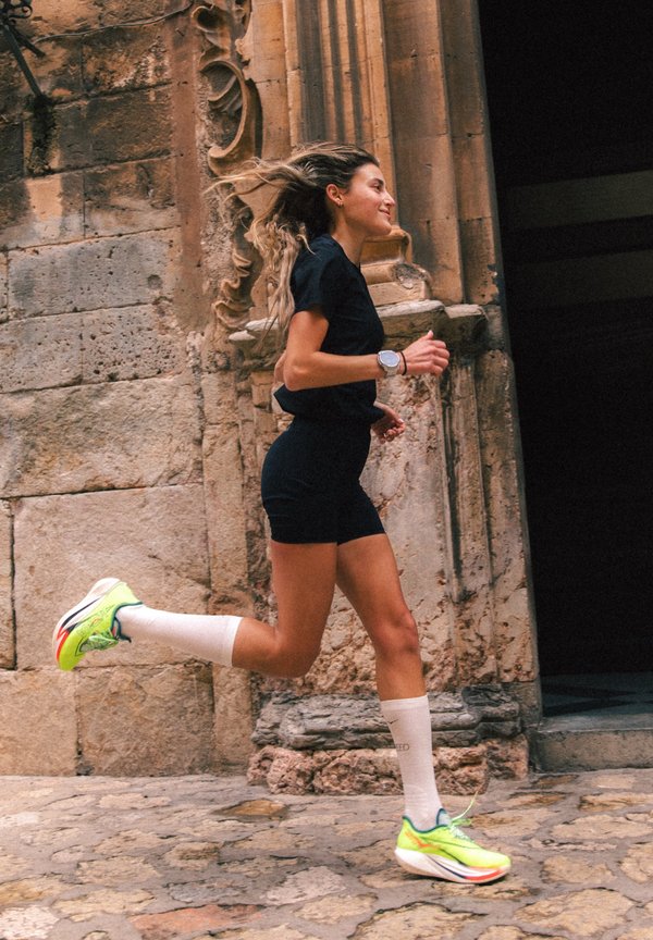 Kobieta w czarnym stroju sportowym i neonowych butach biegowych biegnie obok teksturowanej kamiennej ściany z ozdobnymi elementami architektonicznymi.