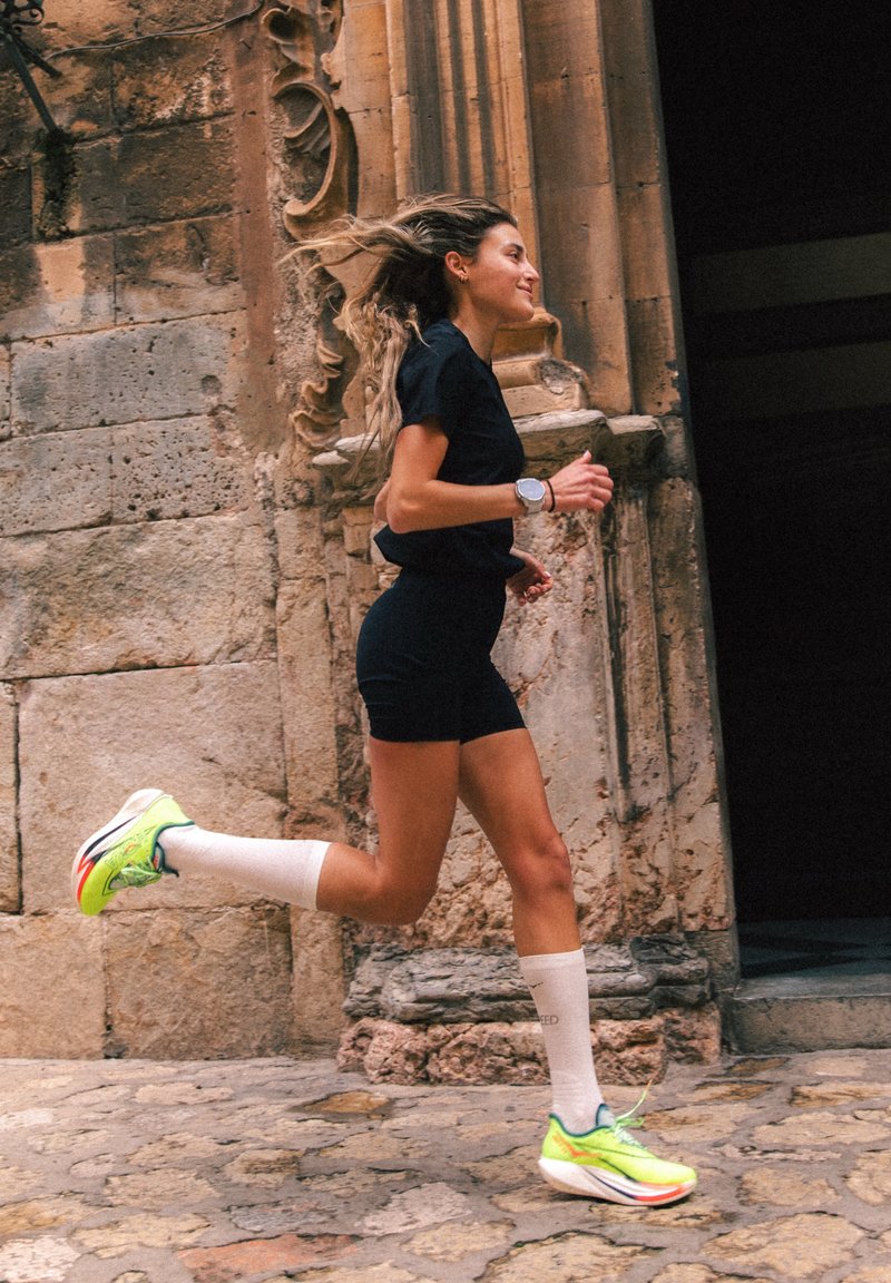 Nő fekete sportöltözetben és neon futócipőben, amint egy faragott kőfal mellett kocog, amely díszes építészeti részletekkel rendelkezik.