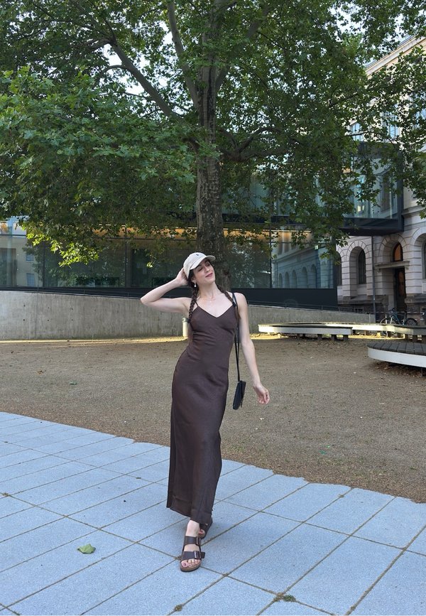 Femme en robe marron, casquette beige et sandales, debout sur une surface pavée près d'un arbre et d'un bâtiment moderne, posant avec une main sur la tête.