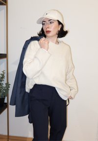 Pull en maille crème avec poignets côtelés, pantalon bleu marine et casquette blanche. Une personne tient une veste foncée sur une épaule. Plante verte en arrière-plan.