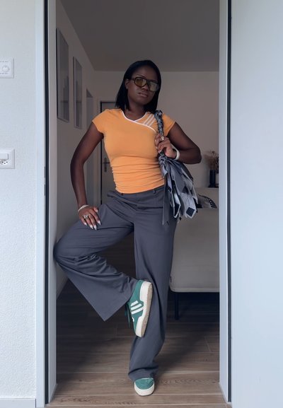 Mujer con top naranja y pantalones grises se apoya en el marco de la puerta con una pierna levantada, sosteniendo un bolso estampado sobre el hombro y llevando gafas de sol.