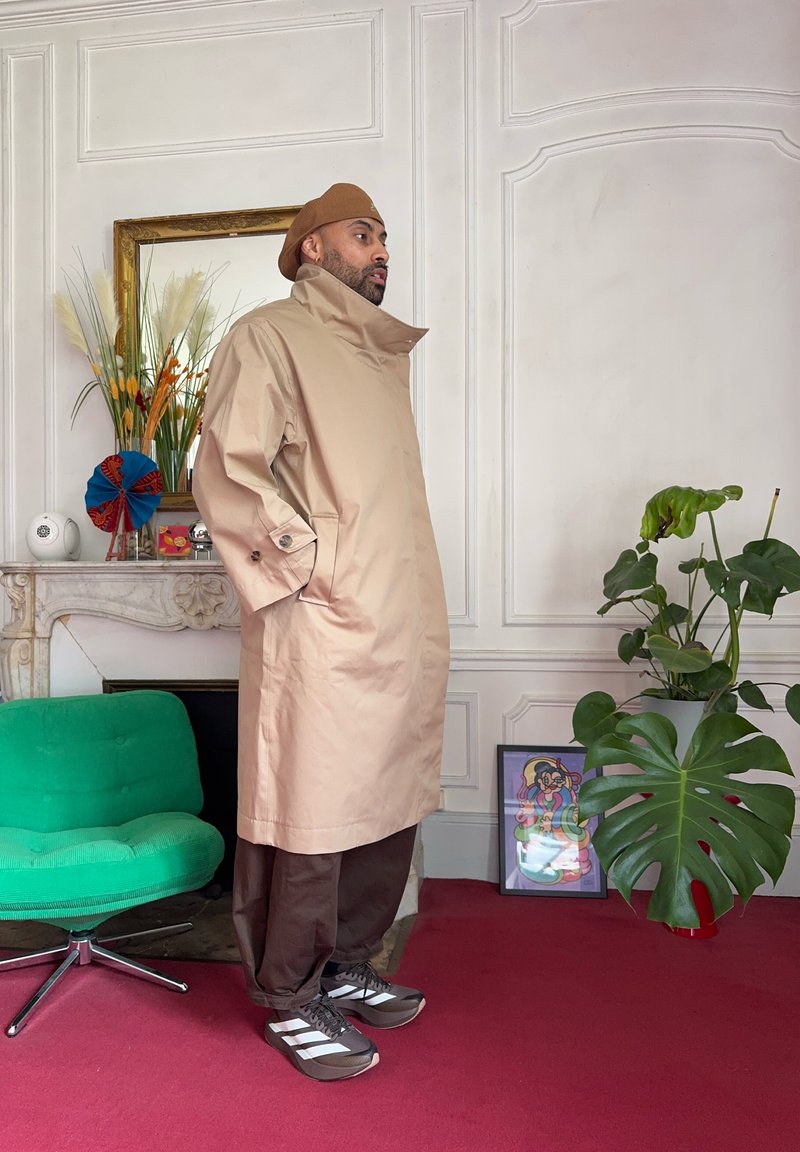 Homme portant un trench-coat beige, un pantalon marron et un chapeau, debout dans une pièce avec une chaise verte, des plantes, une œuvre d'art encadrée colorée et une cheminée décorative.