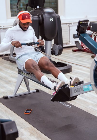 Hombre con camisa blanca y pantalones cortos usando una máquina de remo en el gimnasio, lleva una gorra roja y auriculares, con un teléfono sobre una colchoneta de ejercicio negra cerca.