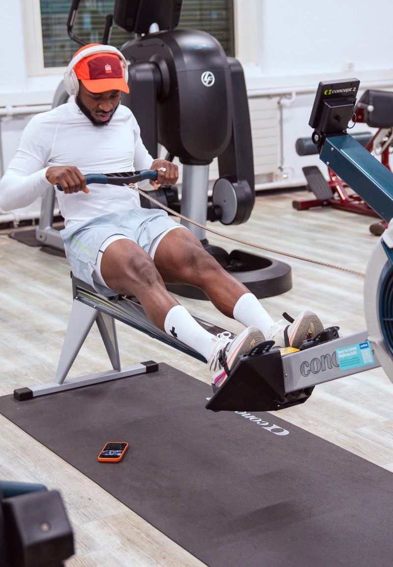 Man in wit T-shirt en korte broek die roeimachine gebruikt in sportschool, met rode pet en koptelefoon, telefoon ligt op een zwarte fitnessmat in de buurt.
