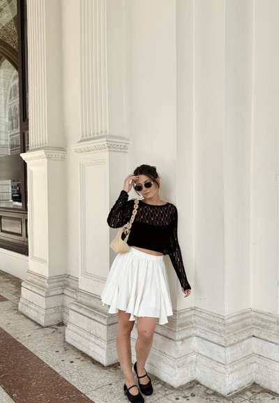 Top de encaje negro de manga larga, falda blanca de corte acampanado, zapatos negros y gafas de sol, con una bolsa de tejido, frente a un fondo de columna blanca texturizada.