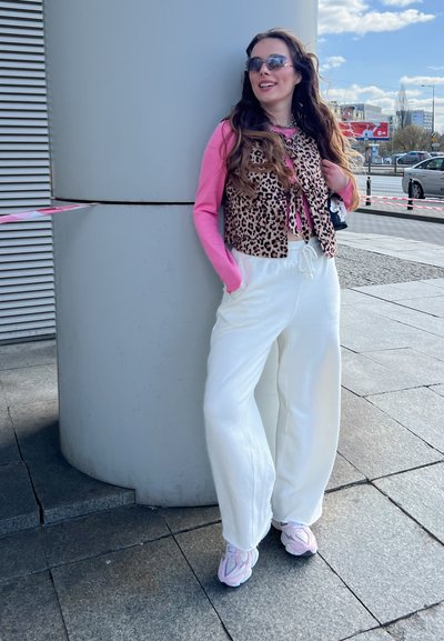 Chaleco de tela de leopardo sobre una camisa de manga larga rosa, combinado con pantalones sueltos blancos y zapatillas pastel. Los accesorios incluyen unas gafas de sol y un bolso pequeño.