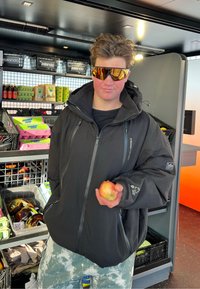 Persona joven con chaqueta negra y gafas de sol reflectantes, sosteniendo una manzana, de pie en una tienda con comestibles en estantes detrás.