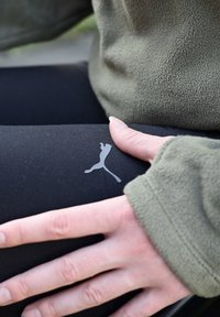 Schwarze Leggings mit einem silbernen Puma-Logo auf dem Oberschenkel, kombiniert mit einem olivgrünen Fleece-Sweatshirt. Nahaufnahme, die eine Hand zeigt, die auf dem Bein ruht.