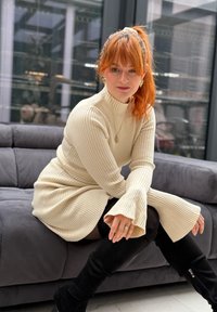 Κοκκινομάλλα γυναίκα με φόρεμα από κρεμ ριμπ πλεκτό και μαύρες μπότες μέχρι το γόνατο, κάθεται σε γκρι καναπέ σε έναν μοντέρνο εσωτερικό χώρο.