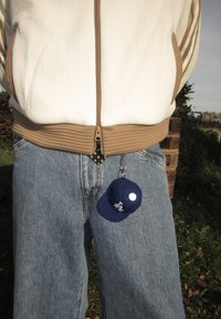 Chaveiro em forma de boné de beisebol mini azul-marinho com detalhe em botão branco, pendurado numa calça de ganga, combinado com um casaco com fecho creme e castanho.