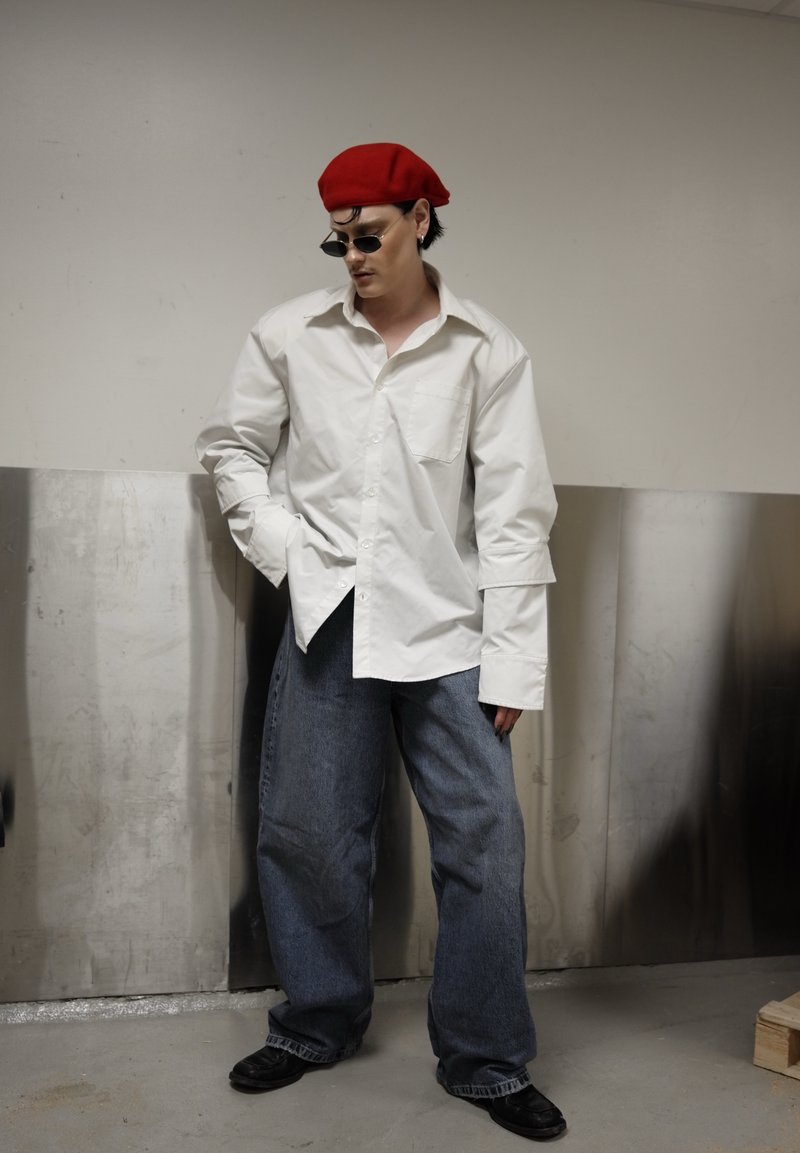 Personne portant une chemise blanche ample, un jean bleu large, un béret rouge, des chaussures noires et de petites lunettes de soleil foncées, debout contre un mur industriel.