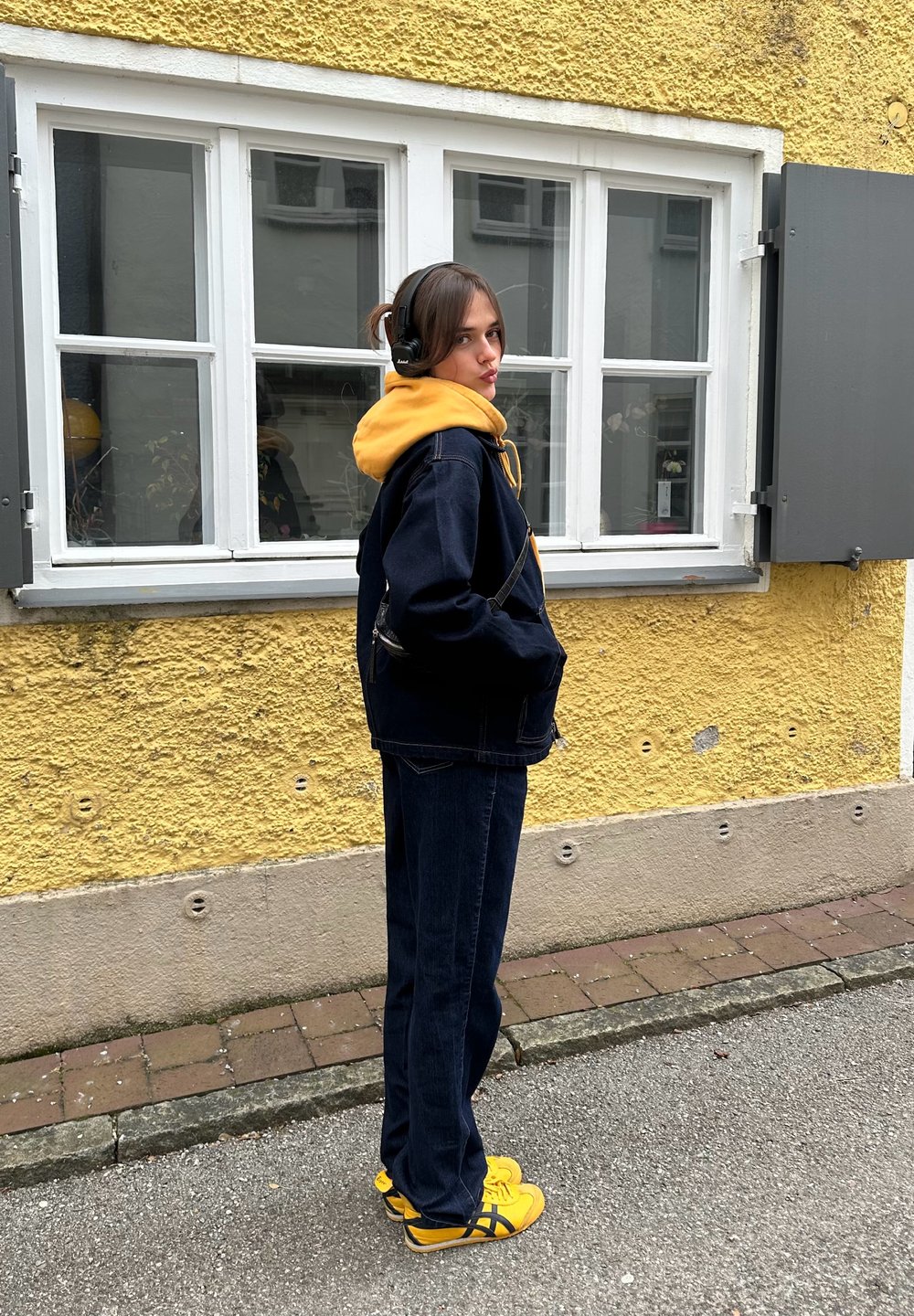 Donkerblauwe spijkerjasje over een gele hoodie, gecombineerd met marineblauwe broek en gele sneakers. Achtergrond bestaat uit een gele muur en witte ramen.