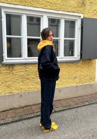 Donkerblauwe spijkerjasje over een gele hoodie, gecombineerd met marineblauwe broek en gele sneakers. Achtergrond bestaat uit een gele muur en witte ramen.