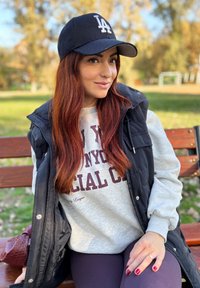 Boné de beisebol preto com logo branco, sweatshirt cinza com texto vermelho, colete preto, leggings escuras e bolsa burgundy em um banco de parque.