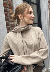 Frau mit hellbraunem Haar im Dutt trägt einen beigen Strickpullover und Schal, goldene abstrakte Ohrringe und hält eine schwarze geflochtene Lederhandtasche neben einer Backsteinmauer.