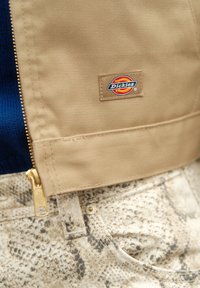 Μπεζ φερμουάρ τζάκετ με λογότυπο, συνδυασμένο με υφασμάτινο μπεζ παντελόνι που διαθέτει σχέδιο φίδι και χρυσές λεπτομέρειες στον φερμουάρ.