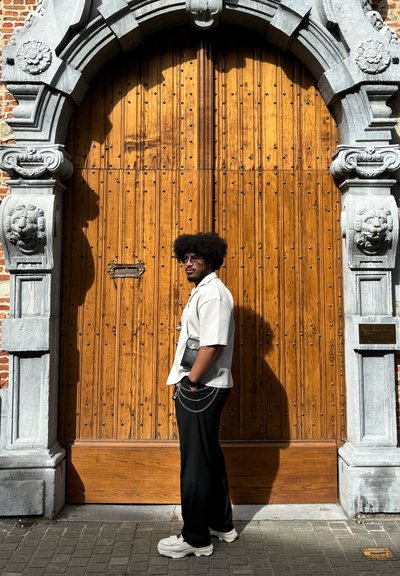 Conjunto informal compuesto por una camiseta de color beige claro, pantalones negros con acentos de cadena y zapatillas blancas, situado frente a una gran puerta de madera.