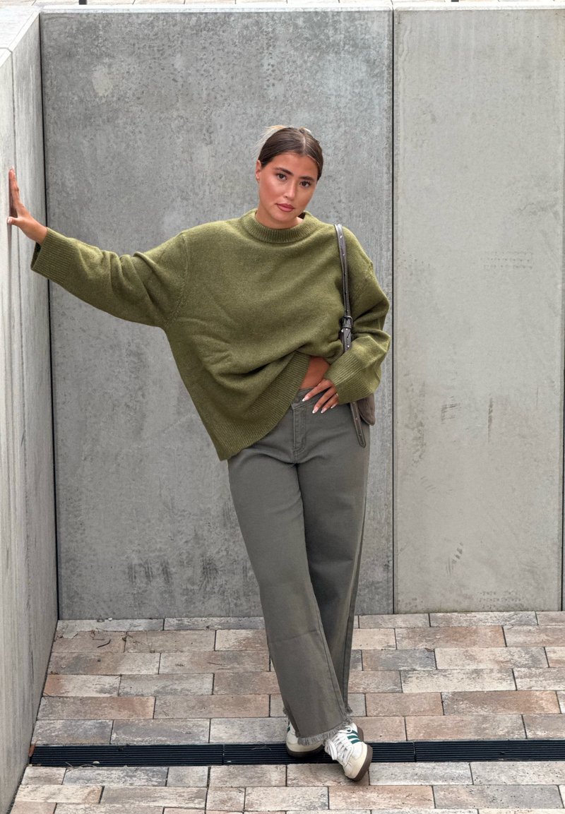 Pulôver de malha verde-oliva oversized com decote redondo, combinado com calças largas cinza-esverdeadas e ténis brancos. O cenário é um fundo de betão.