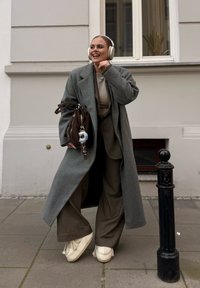Grijze lange wollen jas over een beige top, olijfgroene broek en witte schoenen. Bruine handtas met een sleutelhangerdetail. Achtergrond van een stedelijk trottoir.