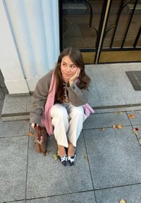 Modella seduta sui gradini, indossa un cappotto grigio, una sciarpa rosa, pantaloni bianchi e décolleté a righe bianche e nere, mentre tiene una borsa marrone.