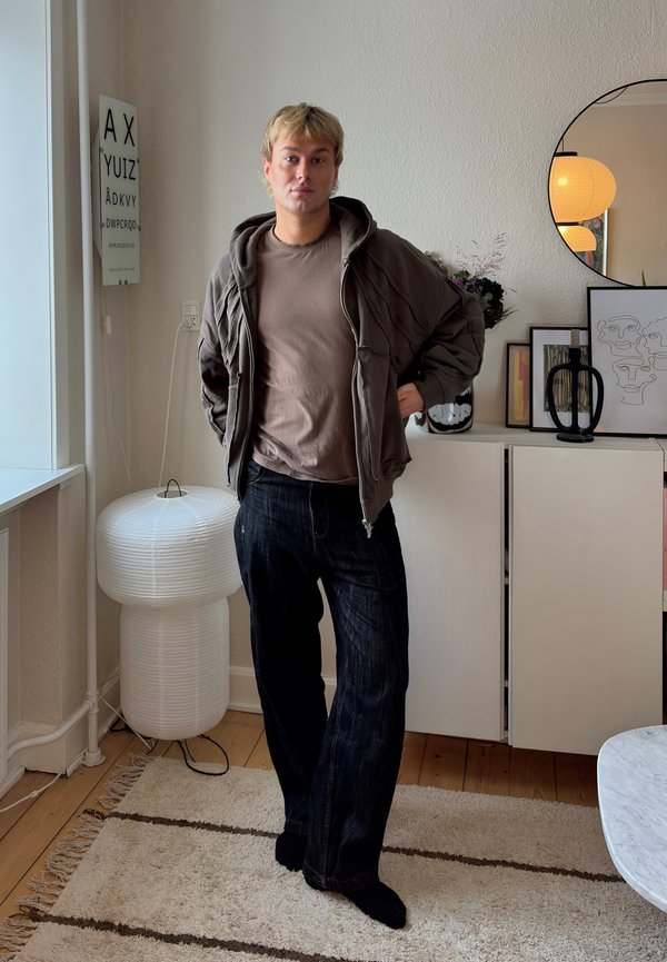 Sweat à capuche gris par-dessus un t-shirt marron, associé à un jean en denim foncé. En arrière-plan se trouvent une lampe circulaire blanche et une œuvre d'art murale décorative.