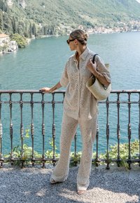 Outfit beige chiaro con design occhiello, camicia a maniche corte con bottoni e pantaloni larghi coordinati. Borsa a tracolla intrecciata e occhiali da sole lisci.