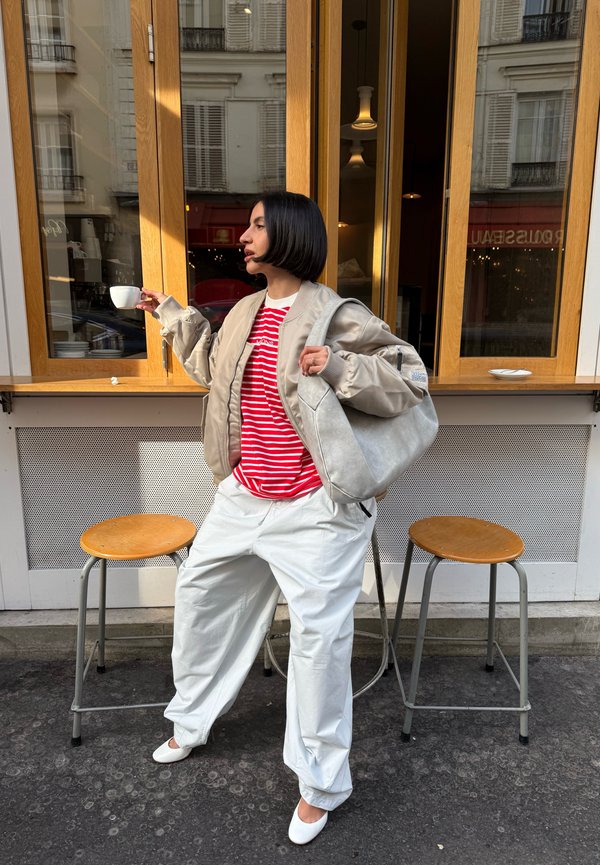 Giacca bomber beige, maglietta a righe rosse e bianche, pantaloni bianchi larghi e tacchi bianchi. Tiene una tazza, in piedi vicino a sgabelli di legno.