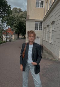 Μαύρο σακάκι πάνω από λευκή μπλούζα, συνδυασμένο με ανοιχτό γαλάζιο τζιν. Μια καφέ τσάντα ώμου συμπληρώνει την εμφάνιση. Αστικό φόντο με δέντρα.