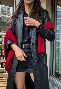 Μαύρο δερμάτινο μακρύ παλτό με τσέπες, συνδυασμένο με μαύρο πουκάμισο με κουμπιά και μαύρες βερμούδες, με έμφαση σε ένα κόκκινο πλεκτό κασκόλ.