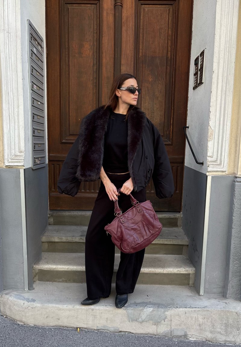 Mujer con chaqueta negra con cuello de piel extragrande y gafas de sol, de pie en los escalones sosteniendo un gran bolso de cuero marrón frente a una puerta de madera.