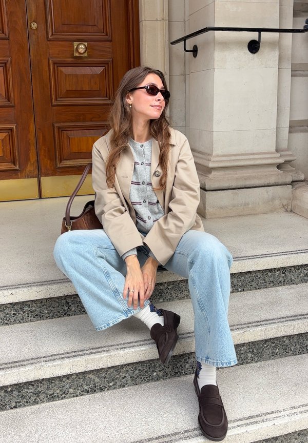 Una mujer con gafas de sol, una chaqueta beige, vaqueros y mocasines está sentada en unos escalones de piedra junto a una puerta de madera.
