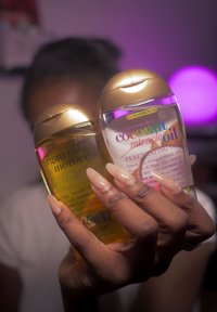 Main avec de longs ongles tenant deux petites bouteilles d'huile capillaire étiquetées « huile d'argan Maroc » et « huile miracle de coco » avec des bouchons dorés.