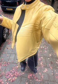 Person iført en gul jakke, svarte bukser og svarte sko som står på en fortau med røde og oransje falne blader.