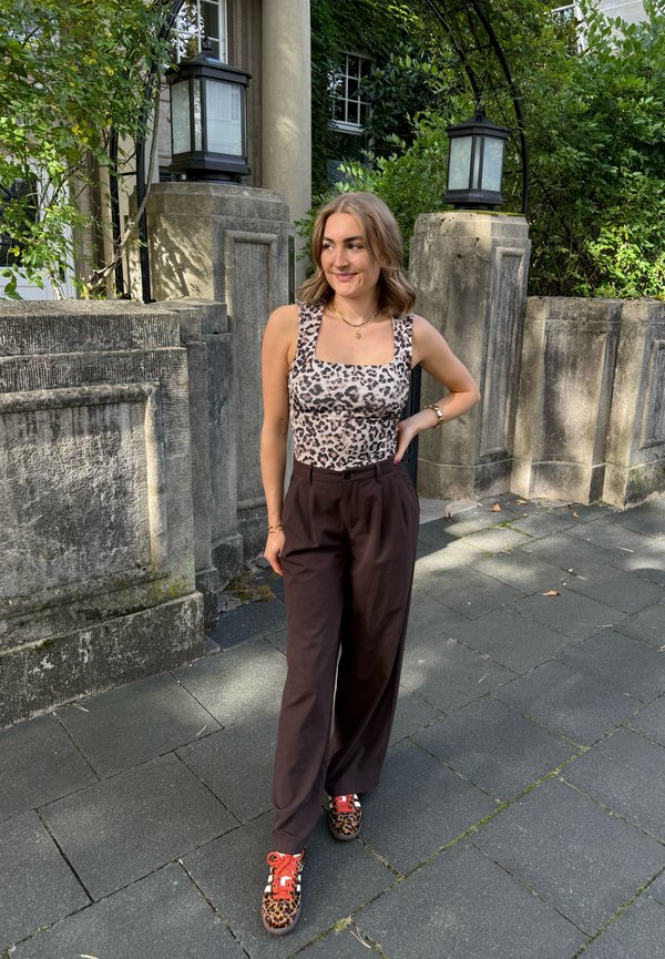 Body léger à imprimé léopard avec décolleté carré, pantalon noir à jambes larges et baskets colorées à imprimé animal. Décor naturel en arrière-plan.