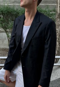 Juodas dvigubas švarkas su išsikirptu apykakle, pagamintas iš lygaus audinio, su dviem priekinėmis kišenėmis ir sagčių akcentais, uždedamas ant balto marškinėlio.