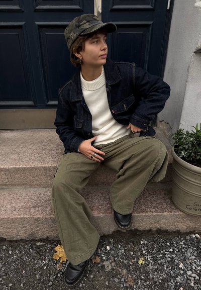 Chaqueta vaquera, suéter crema y pantalones anchos de pierna en oliva. Zapatos negros, gorra de camuflaje, sentado en escaleras de piedra junto a una maceta.